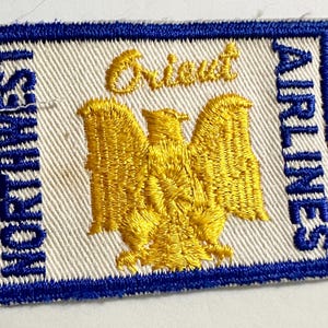 Puede incluir: Un parche de tela blanca con ribete azul y las palabras "Northwest Airlines" y "Orient" bordadas en azul. Un águila bordada en amarillo se encuentra en el centro.