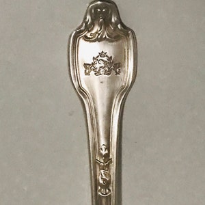 May include: Close-up of the top of a silver spoon with an ornate design. The spoon has a crest with a crown and a letter "S" in the centre.