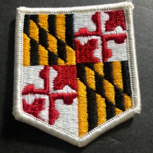 May include: A white embroidered patch with the Maryland state flag. The flag features a black and gold quartered shield with four red crosses on a white background.