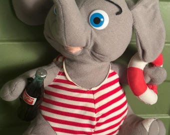 May include: A gray plush elephant toy wearing a red and white striped swimsuit. The elephant has blue eyes and is holding a small Coca-Cola bottle and a red and white life preserver. The toy is on a green surface.