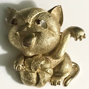 Puede incluir: Una broche de metal dorado de un gato juguetón que sostiene una bola de lana. El gato tiene ojos de pedrería roja y una expresión juguetona.