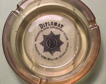 May include: A vintage ashtray with a circular design and a light amber hue. The center features the text "DIPLOMAT" above "RESORTS AND COUNTRY CLUB HOLLYWOOD BY THE SEA, FLORIDA" surrounding a starburst emblem with a "D" in the center.