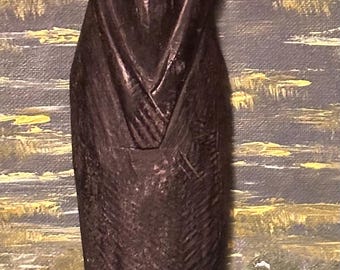 May include: A dark brown wooden sculpture of a standing figure with hands clasped in front. The figure has a detailed face and is wearing a long garment. The base has a cross-hatched design. The background is an abstract painting.