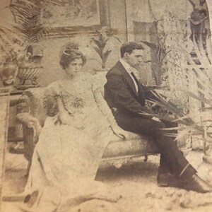 Puede incluir: Fotografía antigua en blanco y negro de un hombre y una mujer sentados en un sofá estampado. La mujer lleva un vestido claro y el hombre un traje. El fondo incluye plantas decorativas y obras de arte, típicas de la época.