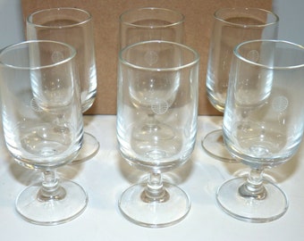 May include: Six clear glass drinking glasses with a white logo on the side. The glasses have a stem and a round base.