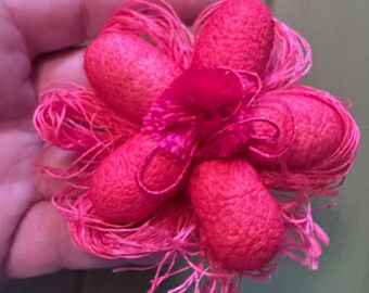 May include: A handmade, vibrant pink flower made of silk cocoons and thread. The flower has five rounded petals, each made from a cocoon, with delicate thread accents. The center is a cluster of pink fabric. The flower is held in a hand.