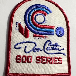 Op de afbeelding: Een witte geborduurde patch met een rood, blauw en wit bowlingpin-ontwerp. De patch heeft een rode rand en de tekst "Don Carter 600 Series" in rood.