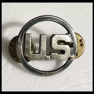 May include: A silver US military pin with a circular design and the letters "US" in the center.