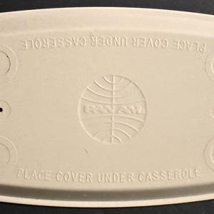 May include: A rectangular, off-white, molded fiber casserole cover featuring the Pan Am globe logo embossed in the center. The text "PLACE COVER UNDER CASSEROLE" is embossed twice along the top and bottom edges, with four circular indentations.