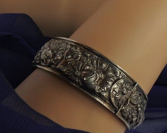 May include: Silver-toned hinged bangle bracelet with detailed floral and insect relief carvings. The bracelet is worn on a wrist, showcasing the intricate design of flowers, leaves, and a bee. The bracelet's clasp is visible.