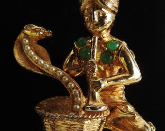 May include: A gold-toned figurine of a musician playing a flute, with a cobra rising from a basket. The cobra has a pearl-like row of beads. The musician and cobra have green gemstone accents. The figurine is set against a black background.