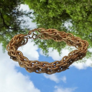 May include: A gold-toned metal chain bracelet featuring a multi-strand woven design with a textured surface. The bracelet is constructed from interconnected oval links that create a thick, braided appearance and includes a circular spring ring clasp.