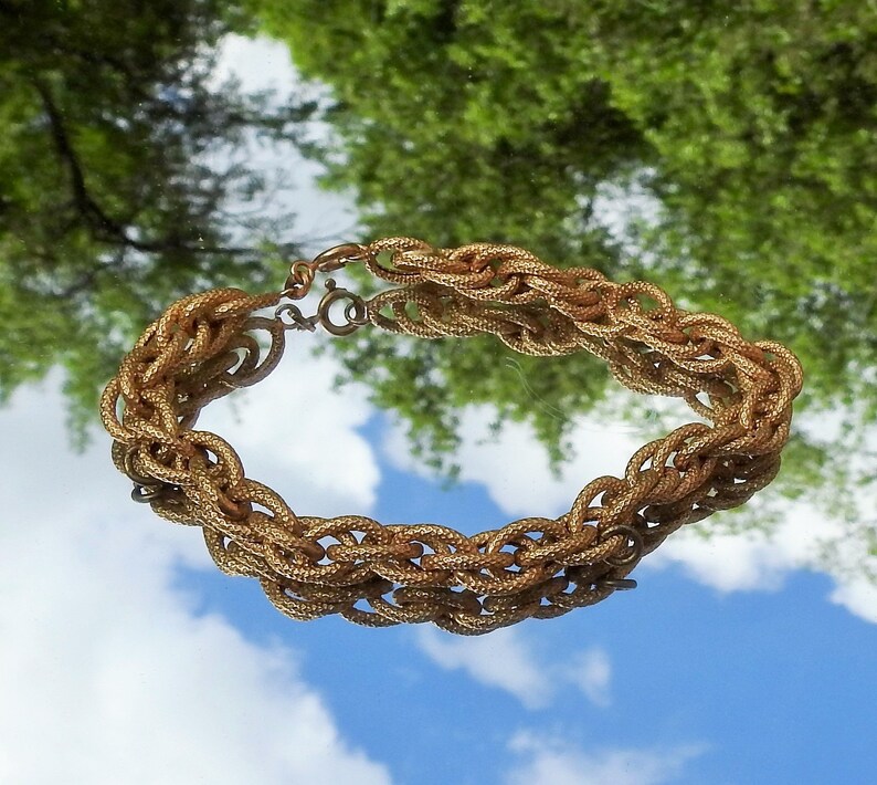 May include: A gold-toned metal chain bracelet featuring a multi-strand woven design with a textured surface. The bracelet is constructed from interconnected oval links that create a thick, braided appearance and includes a circular spring ring clasp.