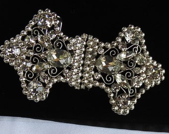 May include: A silver-toned, ornate brooch with a symmetrical design. The brooch is heavily embellished with clear, sparkling rhinestones of various sizes, including large oval and round stones. The design incorporates delicate filigree and scrollwork, creating a glamorous accessory.