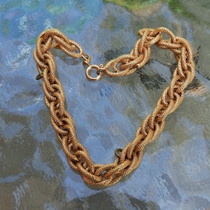May include: A gold-toned chain necklace featuring thick, interlocking oval links with a textured, cross-hatched surface finish. The necklace is secured with a circular spring ring clasp mechanism, creating a continuous, heavy-duty link design.