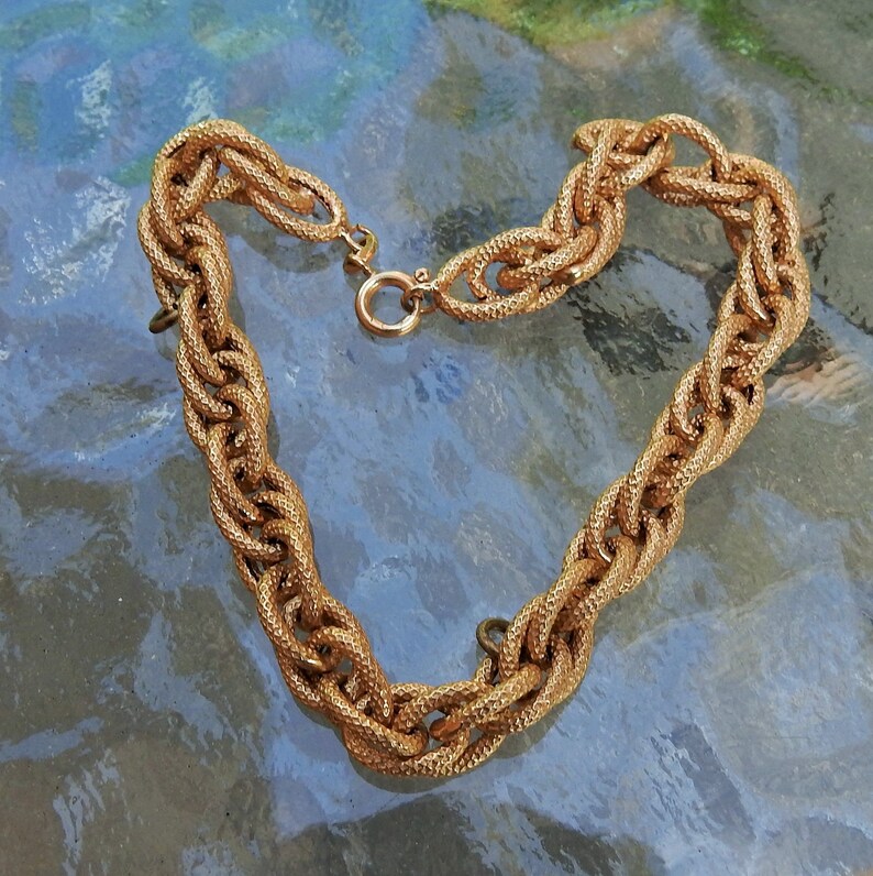 May include: A gold-toned chain necklace featuring thick, interlocking oval links with a textured, cross-hatched surface finish. The necklace is secured with a circular spring ring clasp mechanism, creating a continuous, heavy-duty link design.