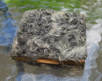May include: A vintage square compact with a textured, fur-like surface in shades of gray and white. The compact has a gold-colored metal frame and clasp, suggesting a classic or antique design. The item is likely a collectible or a unique accessory.