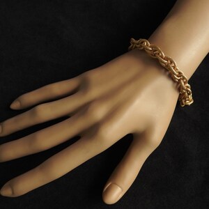 May include: A gold-toned bracelet featuring a thick, braided chain link design with a textured surface. The bracelet is constructed from multiple interlocking metal strands that create a substantial, rope-like appearance around the wrist.