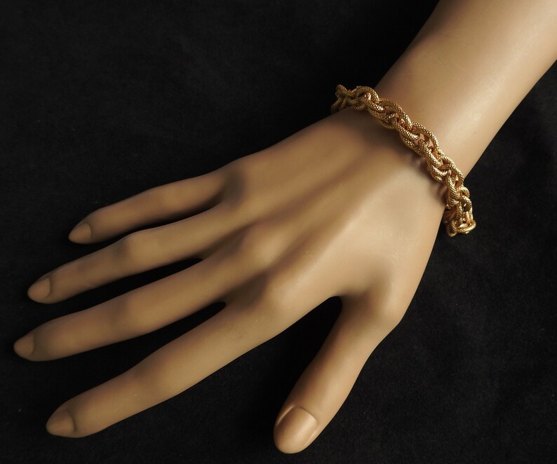 May include: A gold-toned bracelet featuring a thick, braided chain link design with a textured surface. The bracelet is constructed from multiple interlocking metal strands that create a substantial, rope-like appearance around the wrist.
