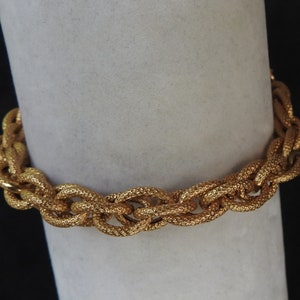 May include: A gold-toned metal bracelet featuring a series of interlocking, textured oval links. The surface of each link has a fine, cross-hatched pattern that creates a matte, granular appearance across the entire length of the chain-style bracelet.
