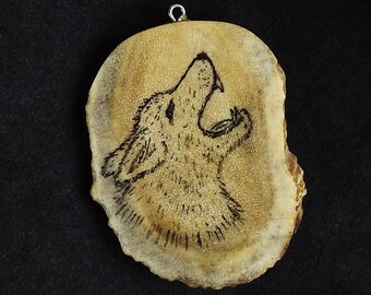 May include: A rustic, oval-shaped pendant with a howling wolf design. The wolf is etched in black on a light brown, textured surface, with a small silver loop at the top for hanging. The background is black.