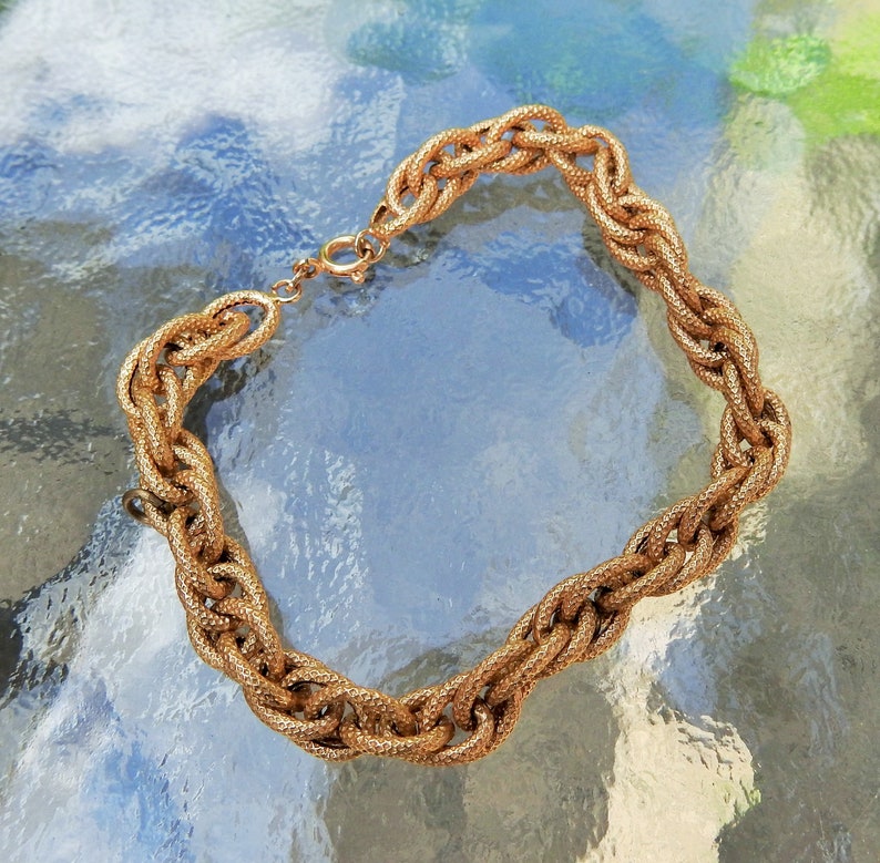 May include: A gold-toned chain bracelet featuring thick, interlocking oval links with a textured, granular surface finish. The bracelet is secured with a small, circular spring-ring clasp mechanism that connects the two ends of the chain.