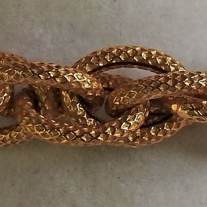 May include: A gold-toned chain necklace featuring thick, interlocking oval links. Each individual link has a textured, cross-hatched surface pattern that creates a repeating diamond-like appearance across the entire length of the metal chain.