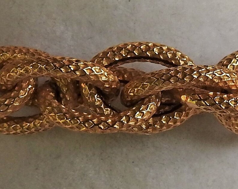 May include: A gold-toned chain necklace featuring thick, interlocking oval links. Each individual link has a textured, cross-hatched surface pattern that creates a repeating diamond-like appearance across the entire length of the metal chain.