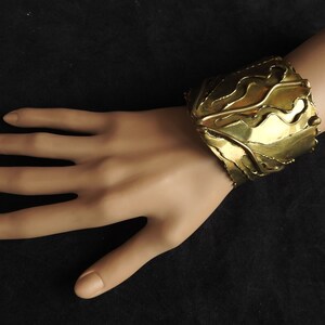 May include: A wide, gold-toned cuff bracelet with an organic, flowing design. The bracelet is worn on a mannequin hand, showcasing its intricate, layered metalwork. The cuff appears to be approximately 2 inches wide.