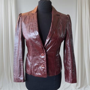 May include: A dark burgundy leather blazer featuring a glossy finish, a notched lapel collar, and long sleeves with slight gathering at the shoulders. The jacket is secured at the front with two matching circular buttons and includes vertical seams.