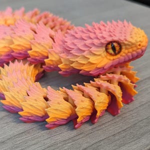 May include: A colorful, articulated snake toy in shades of pink, orange, and yellow. The snake has a detailed, textured design with a realistic eye. The toy is posed on a gray surface.