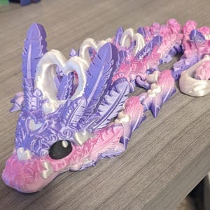 May include: A 3D-printed dragon figurine in shades of purple and pink. The dragon features feather-like details, heart-shaped accents, and a detailed head with a black eye. The figurine is designed with a matching base.