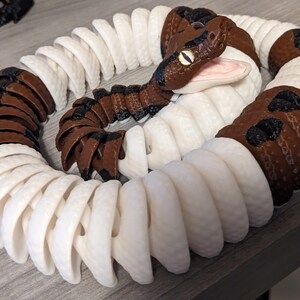 May include: A coiled, articulated snake toy with a brown and white pattern. The snake's head is brown with black accents and yellow eyes. The body alternates between white and brown segments, creating a realistic appearance.