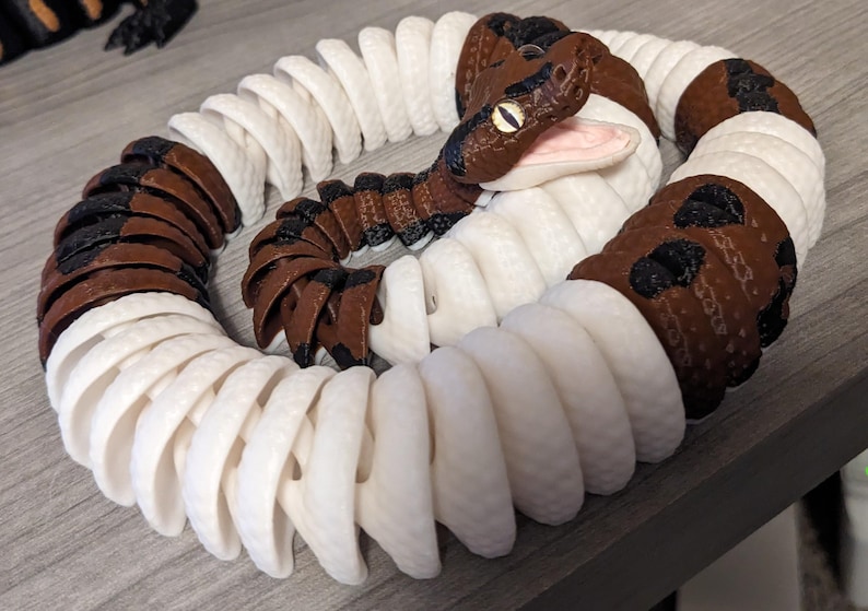May include: A coiled, articulated snake toy with a brown and white pattern. The snake's head is brown with black accents and yellow eyes. The body alternates between white and brown segments, creating a realistic appearance.