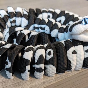 May include: A coiled, black and white snake figurine with a detailed pattern. The snake's head is white with blue eyes. The body is segmented, with alternating black and white sections. The figurine is resting on a light gray surface.
