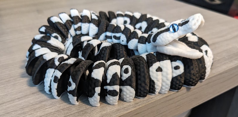 May include: A coiled, black and white snake figurine with a detailed pattern. The snake's head is white with blue eyes. The body is segmented, with alternating black and white sections. The figurine is resting on a light gray surface.