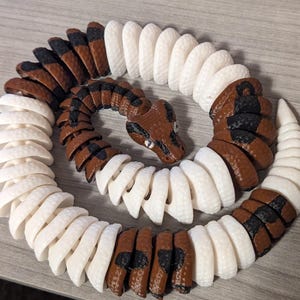 May include: A 3D-printed snake toy in a coiled position. The snake features alternating segments of brown, black, and white. The head is brown with black markings and small, reflective eyes. The toy is designed to resemble a real snake.