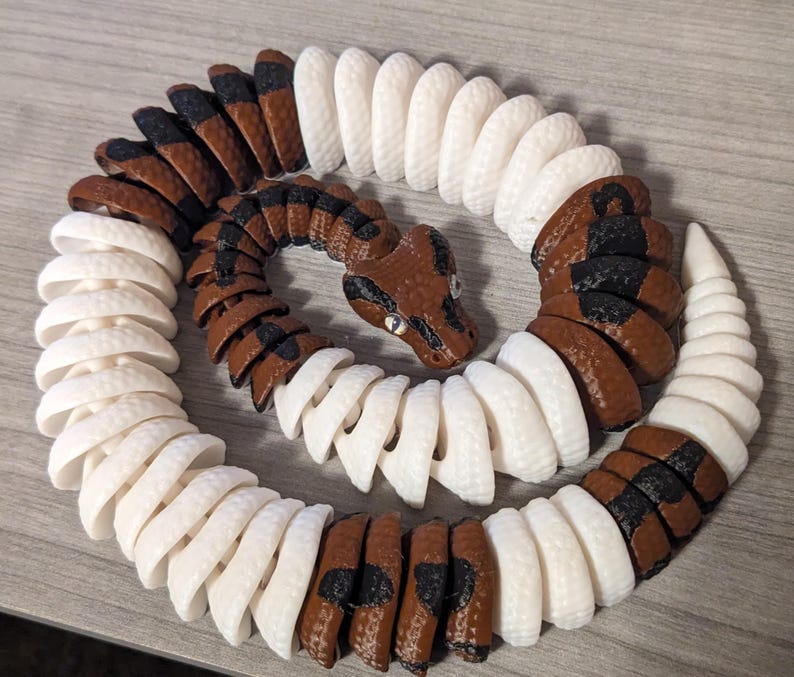 May include: A 3D-printed snake toy in a coiled position. The snake features alternating segments of brown, black, and white. The head is brown with black markings and small, reflective eyes. The toy is designed to resemble a real snake.