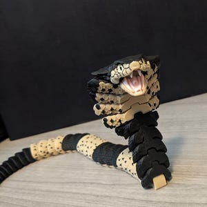 May include: A flexible articulated snake figurine featuring a black and cream-colored segmented body. The cobra-like head is raised with an open mouth revealing teeth and a pink interior. The textured scales alternate between black and cream patterns.