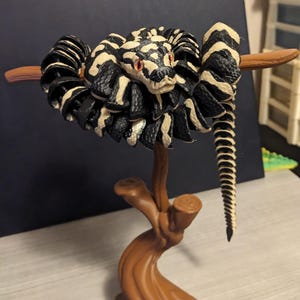 May include: A detailed sculpture of a snake coiled around a brown branch. The snake features a black and cream pattern, with red eyes. The sculpture is mounted on a brown, stylized tree trunk, creating a unique decorative piece.