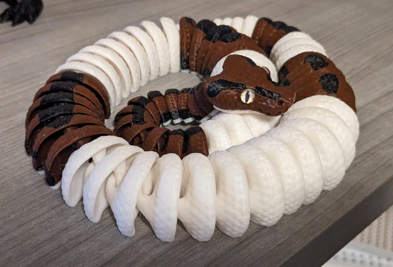 May include: A 3D-printed articulated snake toy in a coiled position. The snake features a brown and white color scheme with black spots. The head is brown with a detailed eye. The toy is designed to be flexible and poseable.