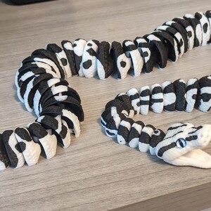 May include: A segmented snake toy in black and white, with a detailed head and open mouth. The toy is made of multiple connected pieces, allowing it to be posed. The snake is lying on a light brown surface.