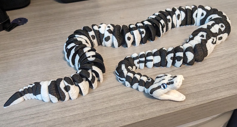May include: A segmented snake toy in black and white, with a detailed head and open mouth. The toy is made of multiple connected pieces, allowing it to be posed. The snake is lying on a light brown surface.