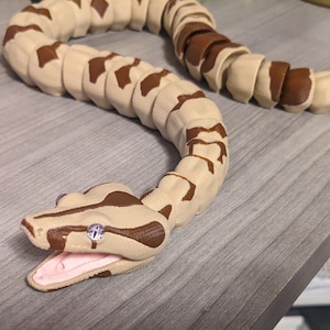 May include: An articulated toy snake featuring a segmented body with a tan and brown patterned design. The snake has a detailed head with an open mouth showing a light pink interior and a single visible eye with a vertical pupil and silver iris.