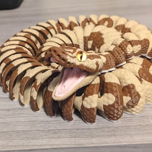 May include: A coiled snake figurine with a detailed design. The snake features a brown and cream pattern, an open mouth revealing a pink interior, and a yellow eye. The figurine is likely made of a durable material.