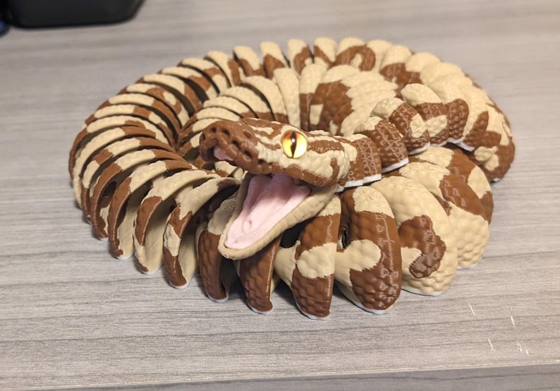 May include: A coiled snake figurine with a detailed design. The snake features a brown and cream pattern, an open mouth revealing a pink interior, and a yellow eye. The figurine is likely made of a durable material.