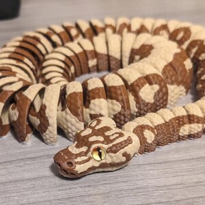 May include: A coiled, segmented snake toy in shades of brown and cream. The snake's head is detailed with a brown pattern and a yellow eye. The toy is designed to appear realistic and is positioned on a light gray surface.