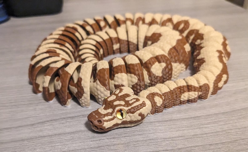 May include: A coiled, segmented snake toy in shades of brown and cream. The snake's head is detailed with a brown pattern and a yellow eye. The toy is designed to appear realistic and is positioned on a light gray surface.