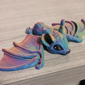 May include: A 3D-printed, colorful bat figurine with a blue head, large black eyes, and rainbow-colored wings. The bat is lying down on a wooden surface, showcasing its detailed design and vibrant color scheme.