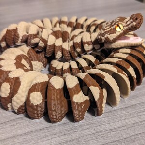 May include: A 3D-printed articulated snake toy in shades of brown and cream. The snake's body is segmented with a repeating pattern. The head is detailed with a realistic eye and open mouth, showcasing a pink interior.
